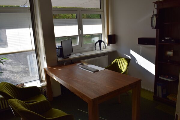 Modernes Büro mit Holztisch, grünen Sesseln, Computer und großem Fenster im Sonnenlicht.
