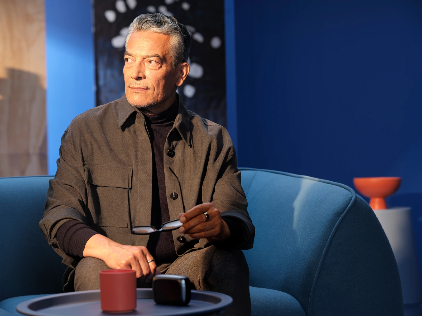 Älterer Mann sitzt nachdenklich auf blauem Sofa mit Brille in moderner Wohnzimmerumgebung.